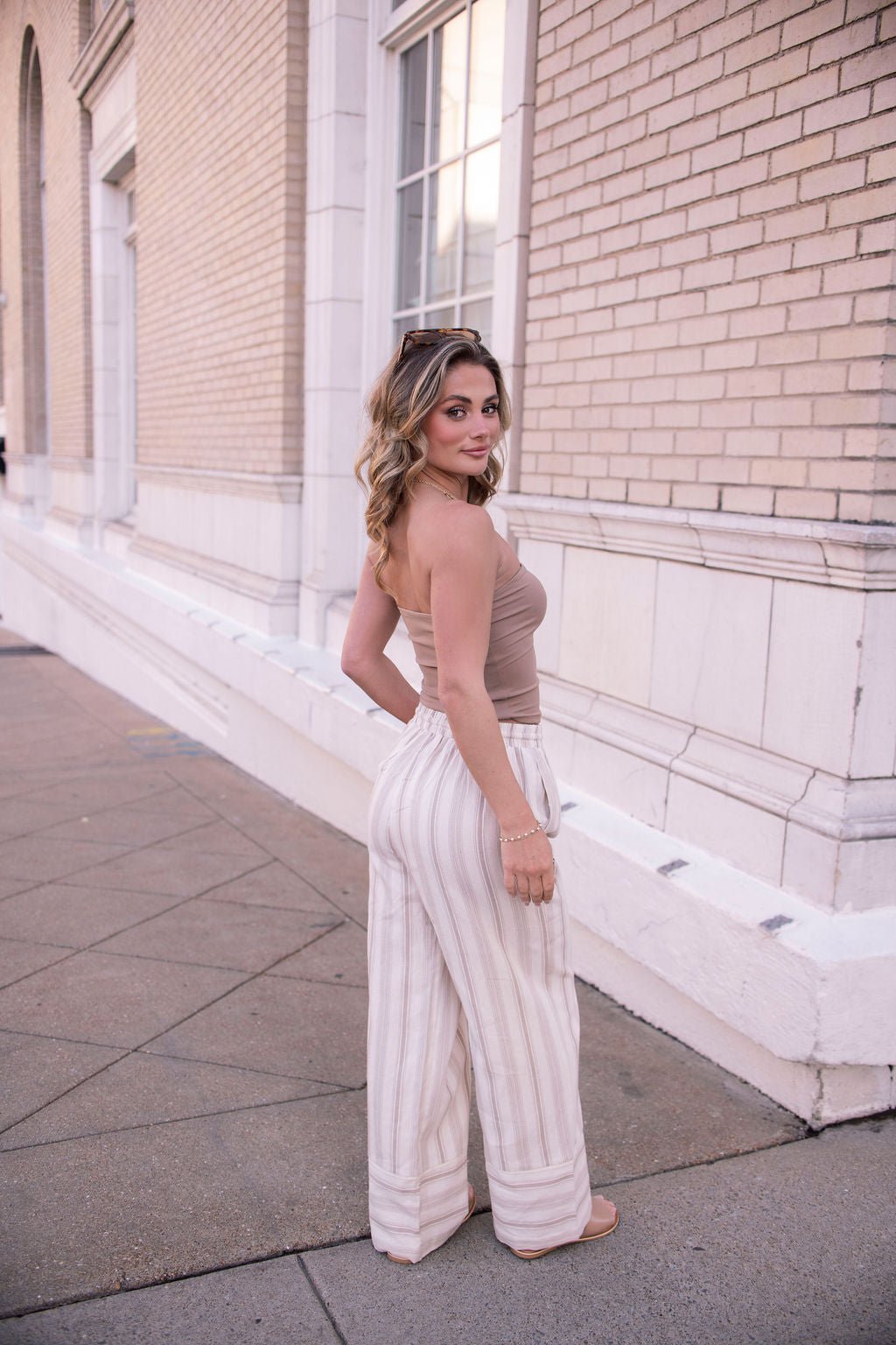 A woman in a strapless top and Mocha Striped Palazzo Pants | Pre Order smiles as she looks back over her shoulder on the sidewalk.