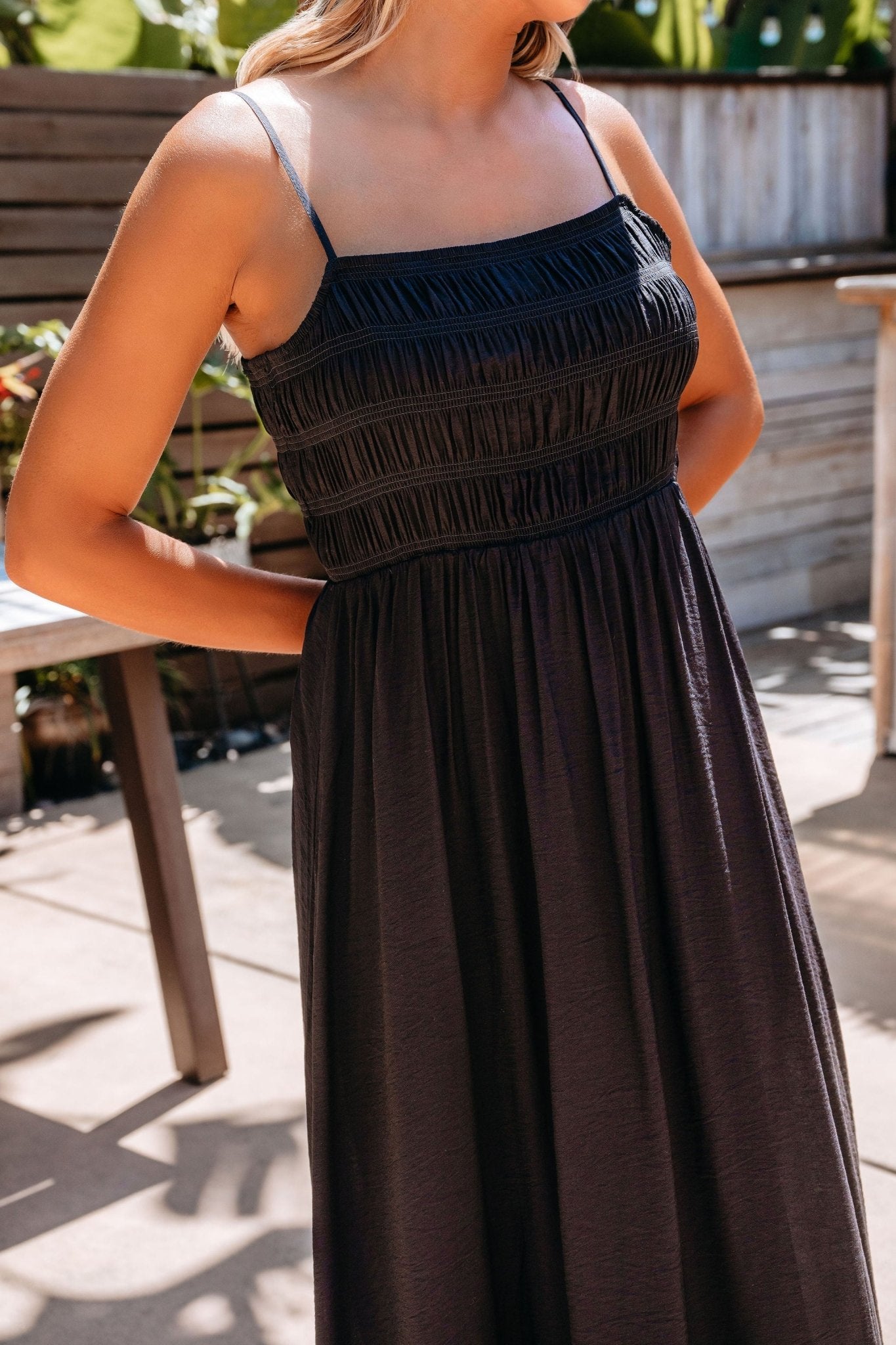 Woman wears the Moonlight Hour Black Ruched Maxi Dress with thin straps and pockets, standing outdoors.