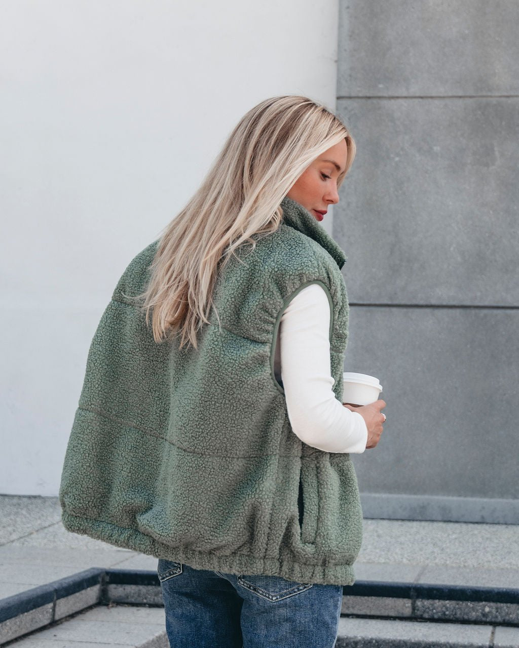 A woman with long blonde hair stands outdoors in the Mountainside Zip Up Fleece Vest - Sage, holding a coffee cup.