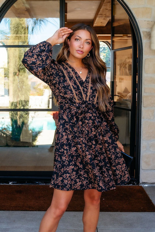 Woman in the Multi Black Floral Print Mini Dress and tan heels poses outside, touching her hair.