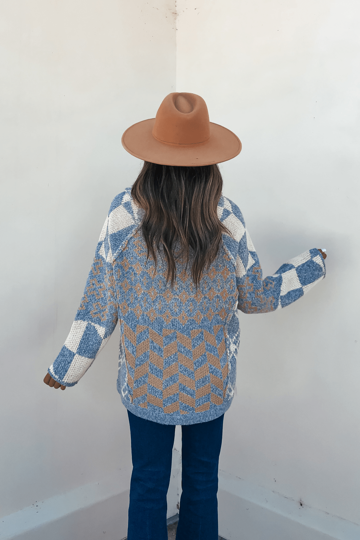 Woman in a Multi Blue Contrast Button Down Sweater Cardigan and brown hat, back to camera against a white wall.