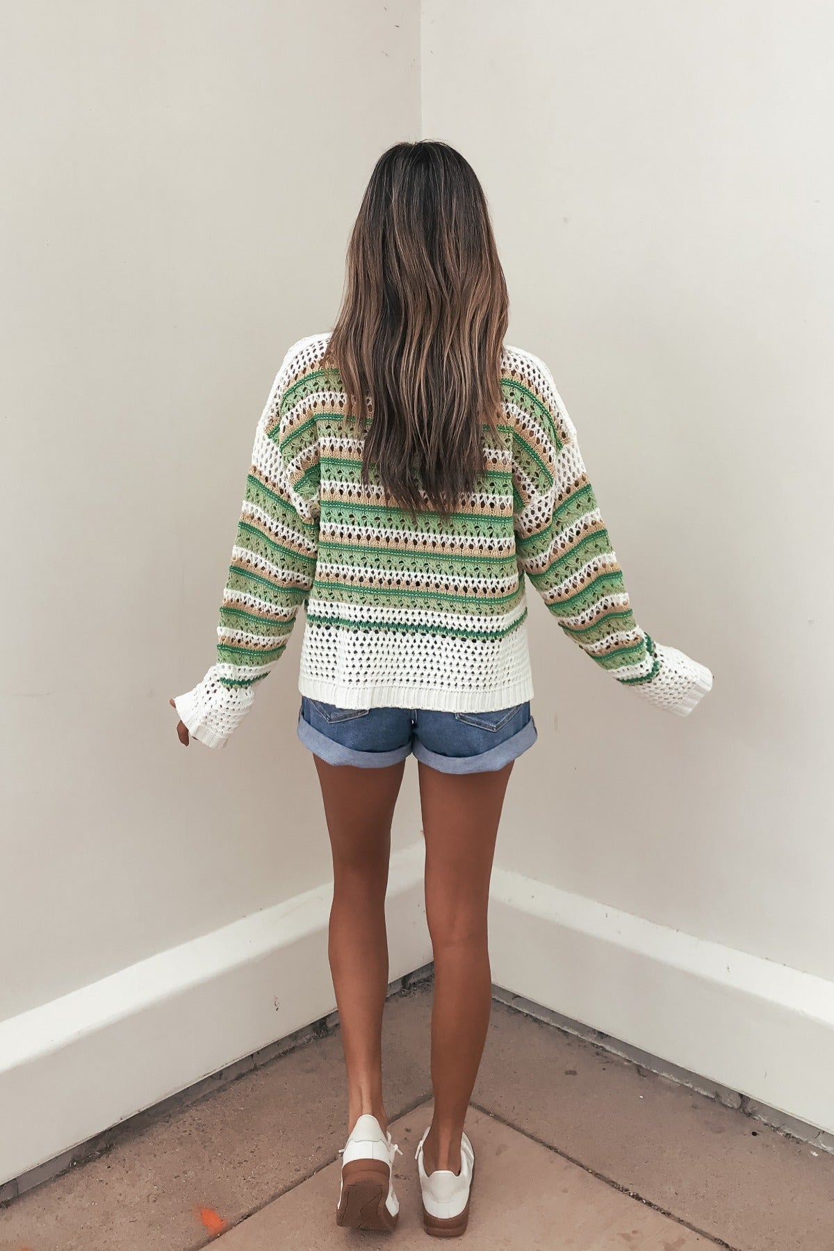 Woman with long hair faces a white wall, wearing a Multi Green Striped Boatneck Sweater, denim shorts, and white shoes.