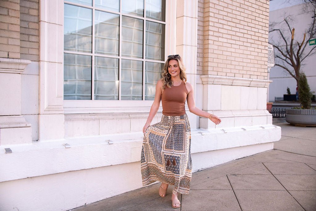 Woman wearing the Multi Olive Print Smocked Maxi Skirt - FINAL SALE smiles while walking by a sunny window.
