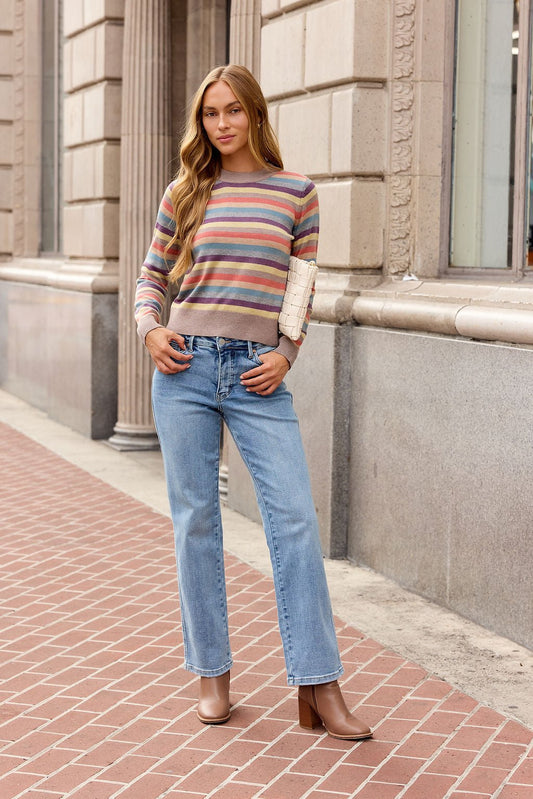 Woman wearing a Multi Stripe Ribbed Sweater, blue jeans, and brown boots stands on a brick sidewalk holding a small clutch.