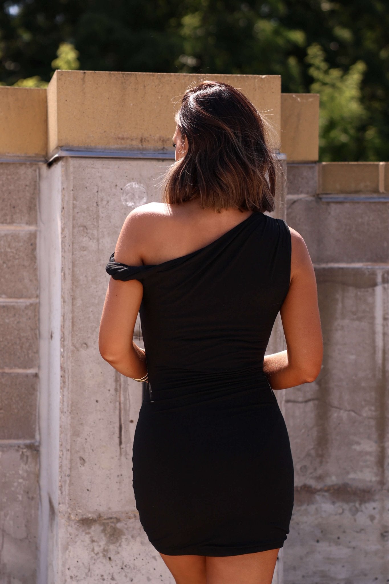A woman stands outdoors with her back to the camera, wearing the Natalia Black Asymmetrical Mini Dress - FINAL SALE.