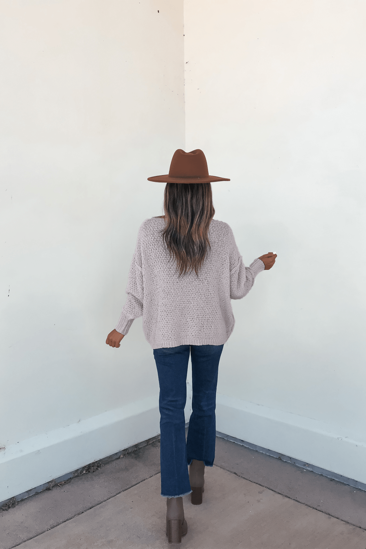 Woman stands in a corner facing the wall, wearing a Natasha Chunky Cable Knit Sweater in grey, brown hat, jeans, and boots.