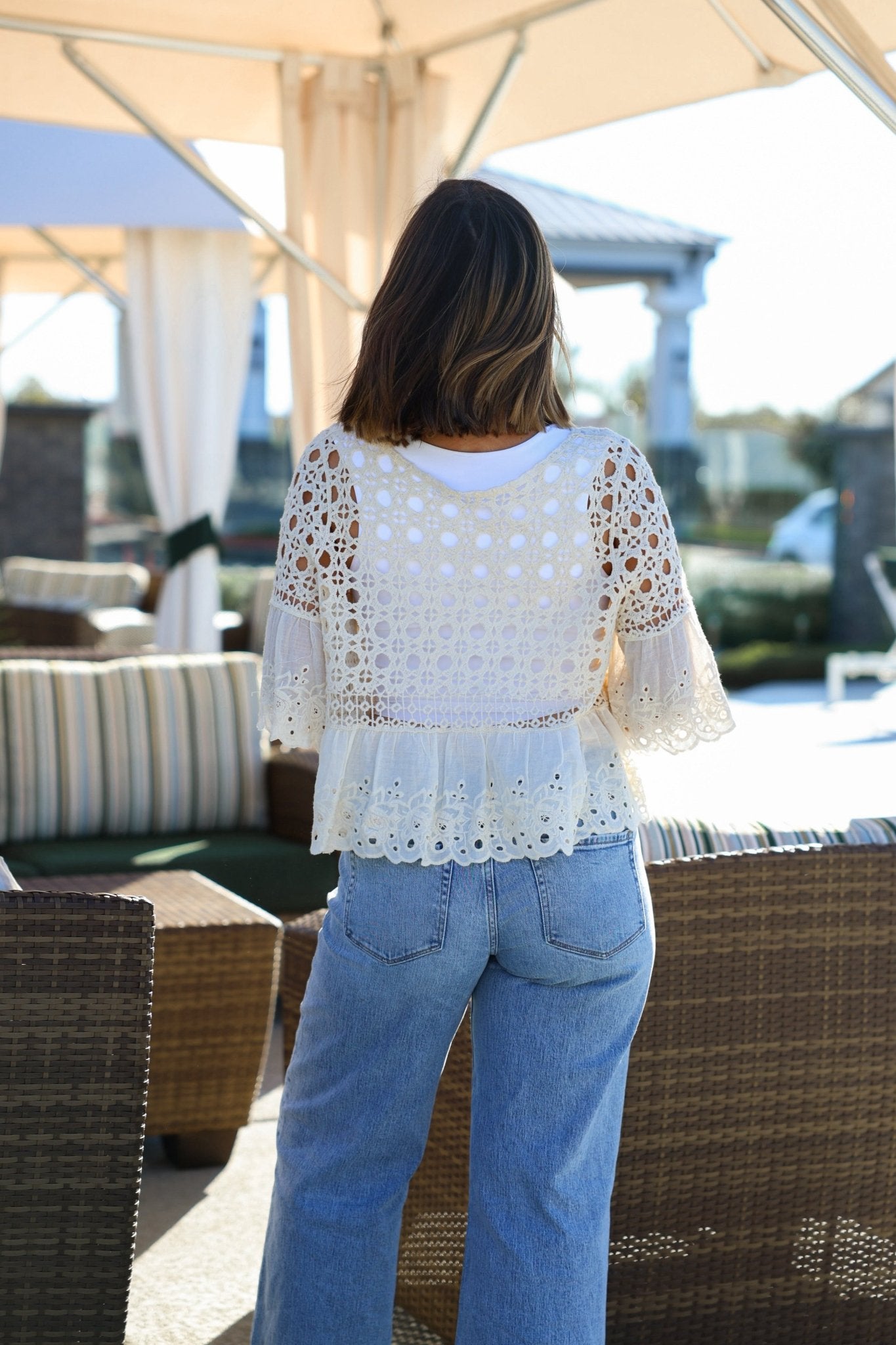 A woman with brown hair in blue jeans stands outdoors, her back to the camera, wearing a Natural Crochet Floral Eyelet Top.