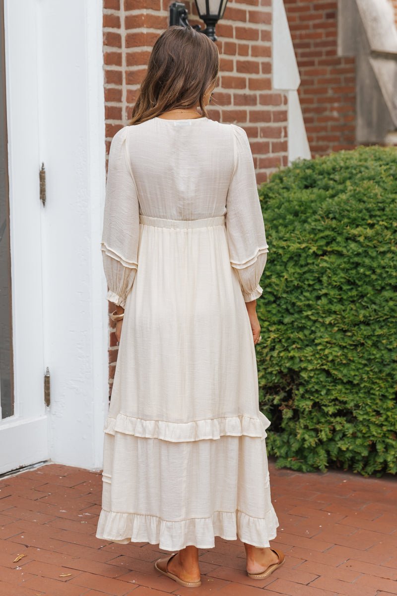 A woman wears the Natural Lace Embroidered Tiered Maxi Dress while standing on a brick path near a green bush and white door.