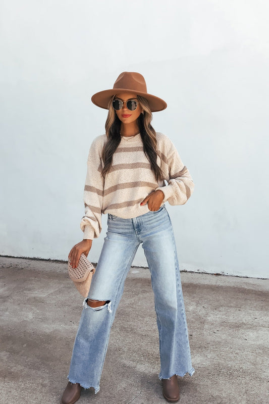 Woman in a brown hat, Natural & Mocha Striped Sweater, ripped jeans, and boots stands outdoors with a woven bag.