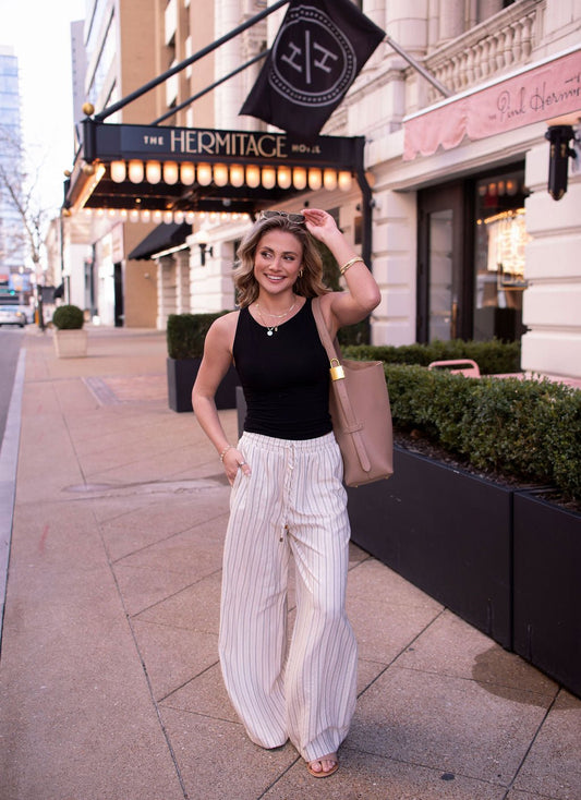 A woman in Natural Striped Linen Drawstring Pants walks on a city sidewalk, smiling and carrying a tote bag.