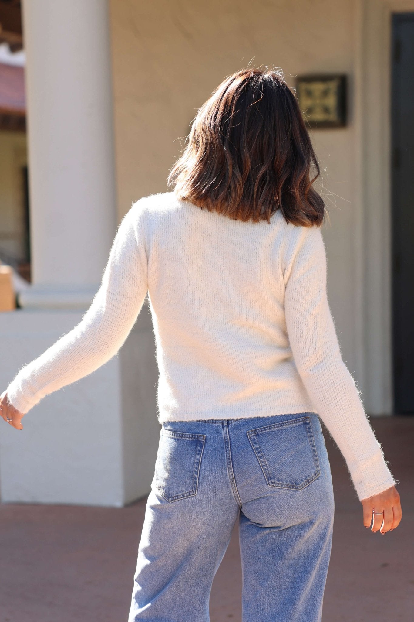 A woman with shoulder-length brown hair in a white sweater and blue jeans layers with the Natural Tie Front Cardigan - FINAL SALE.