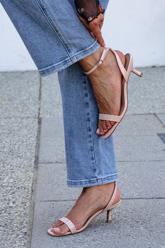 Person in blue jeans adjusts the ankle strap on Nude Strappy Kitten Heels while outdoors.