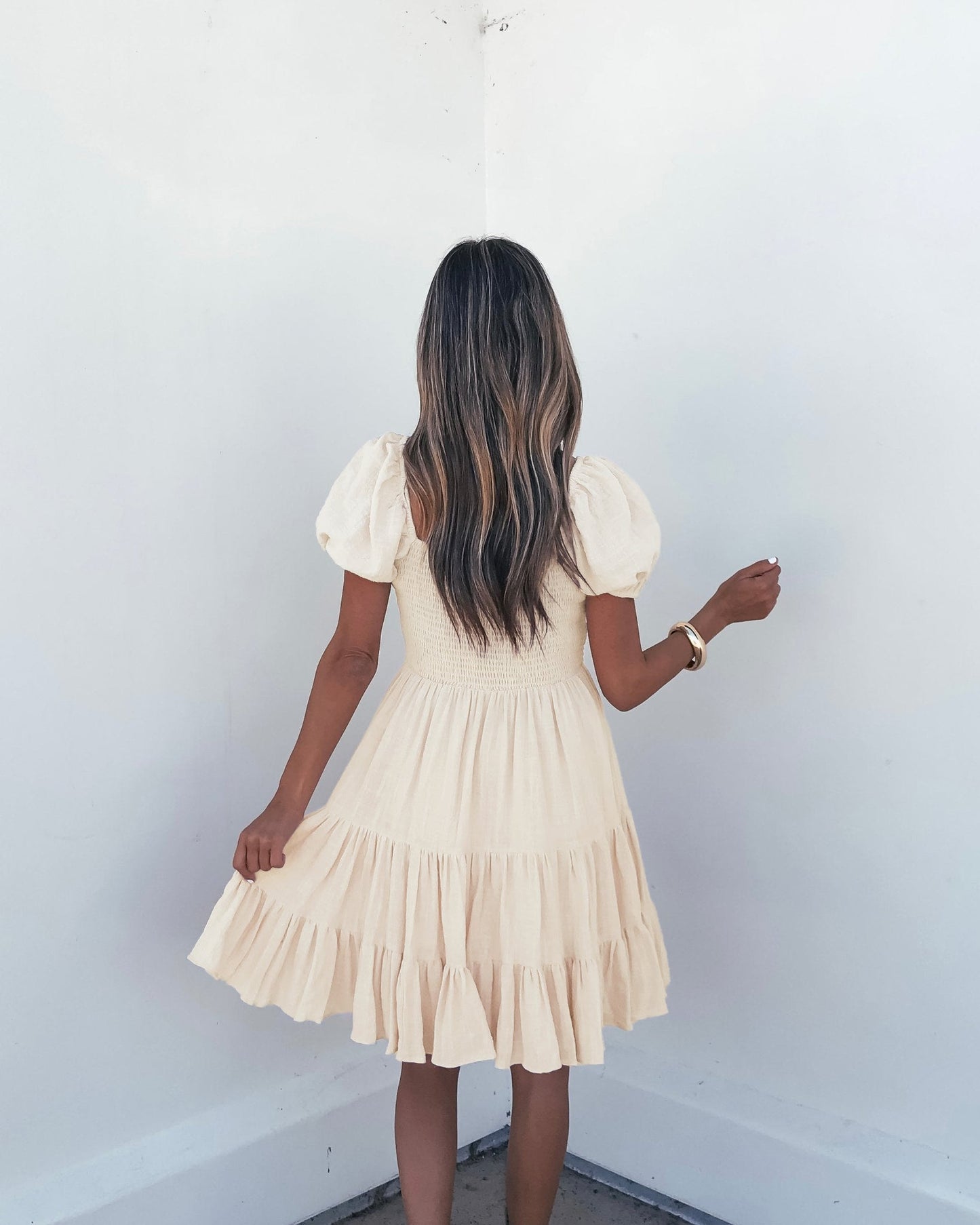 A woman with long wavy hair wears the Oatmeal Puff Sleeve Tiered Mini Dress - FINAL SALE, holding its skirt by a white wall.