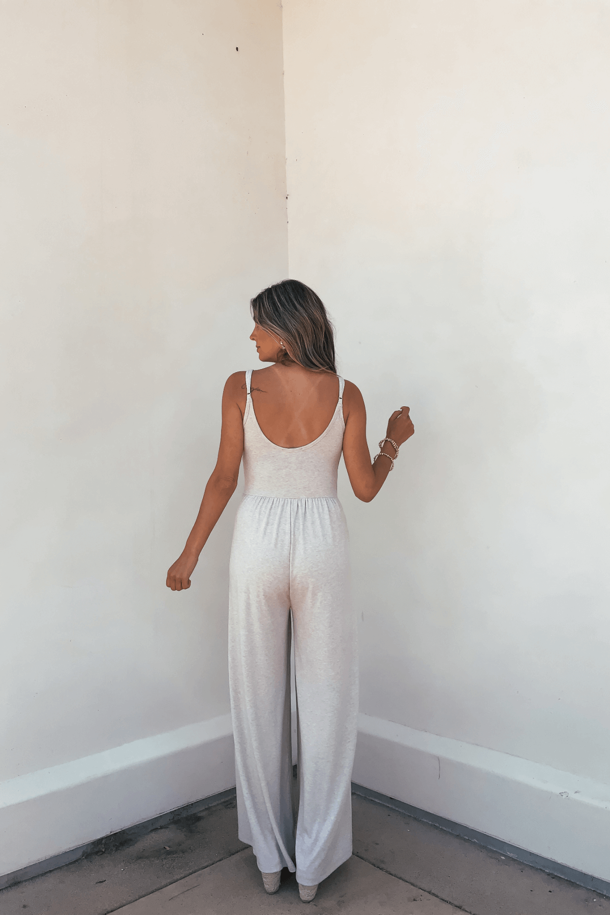 Woman facing away in Oatmeal Scoop Neck Wide Leg Jumpsuit - FINAL SALE, against a plain off-white wall.