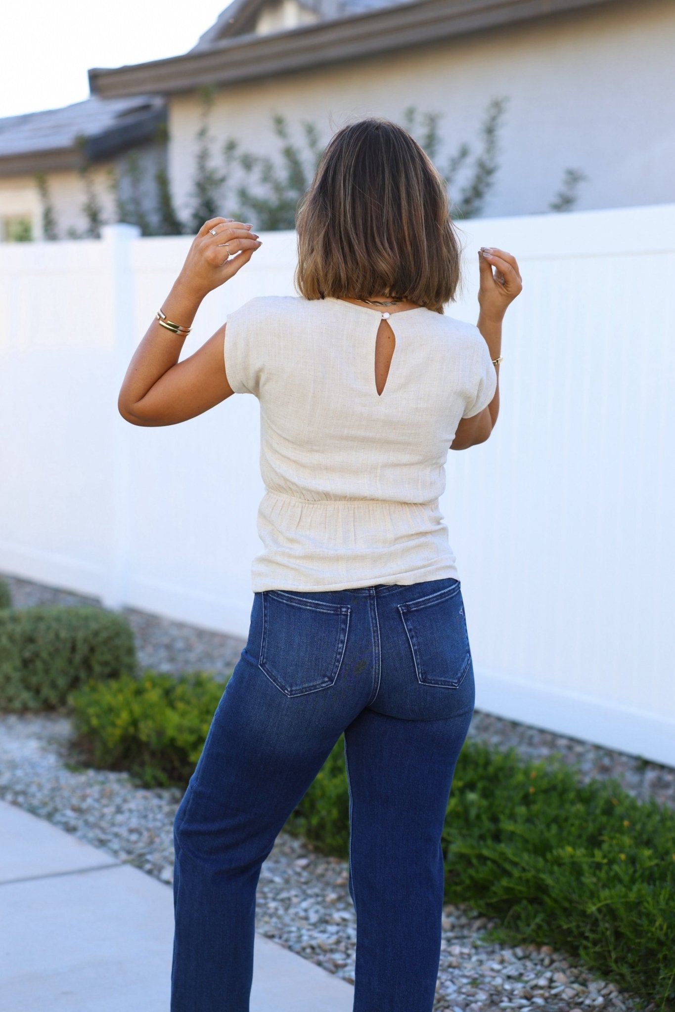 A woman in an Oatmeal Shirred Linen Top and blue jeans stands outdoors with her back to the camera.