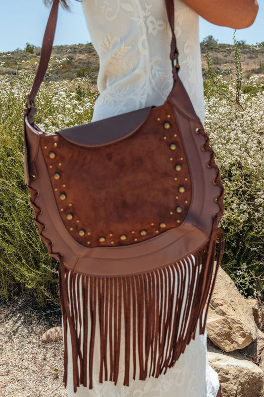 A woman in a white dress holds the Outlaw Fringe Shoulder Bag - Brown, standing outdoors among greenery.