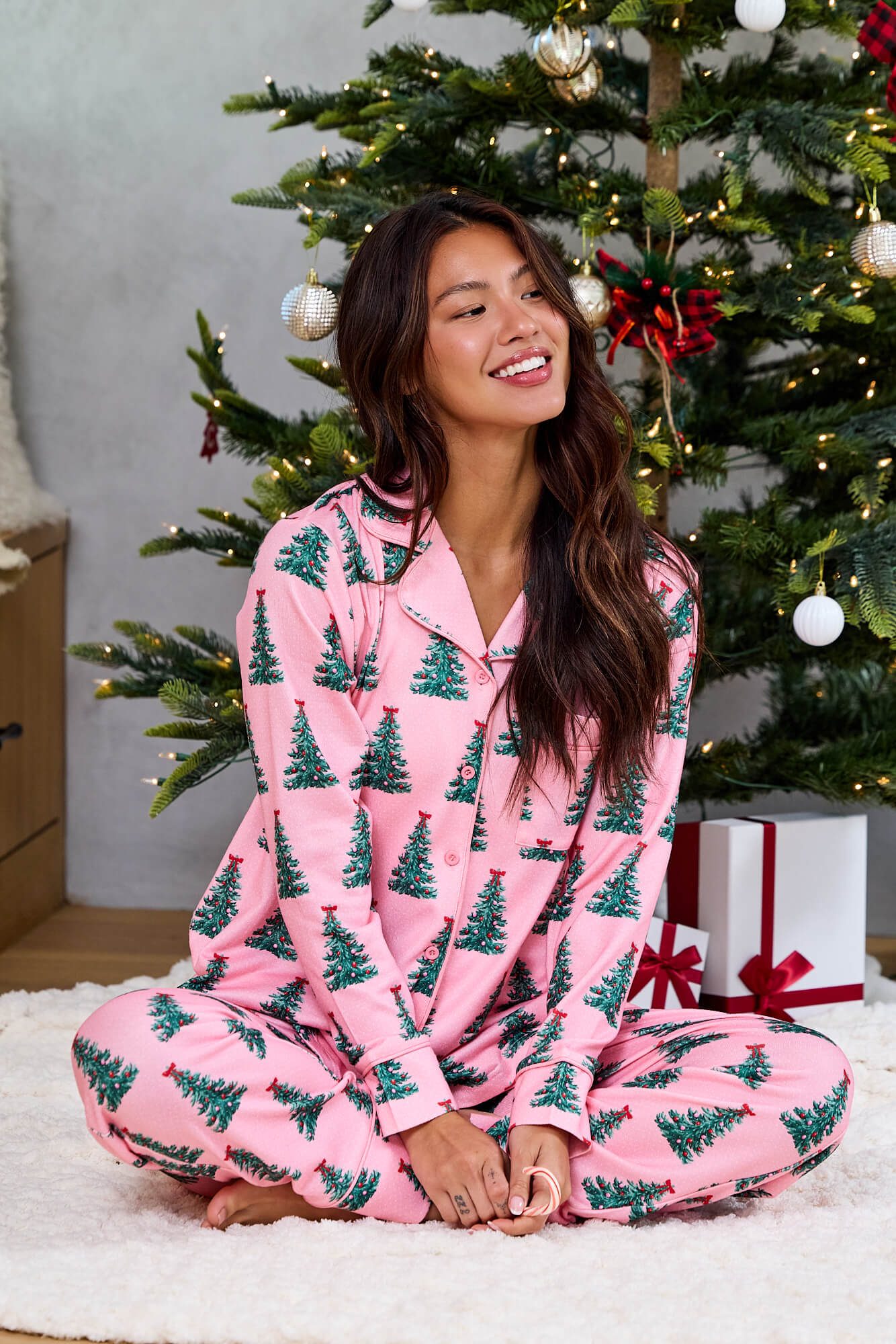 A woman in the Pink Christmas Tree Print Pajama Set - FINAL SALE smiles on a rug by a decorated tree and festive presents.