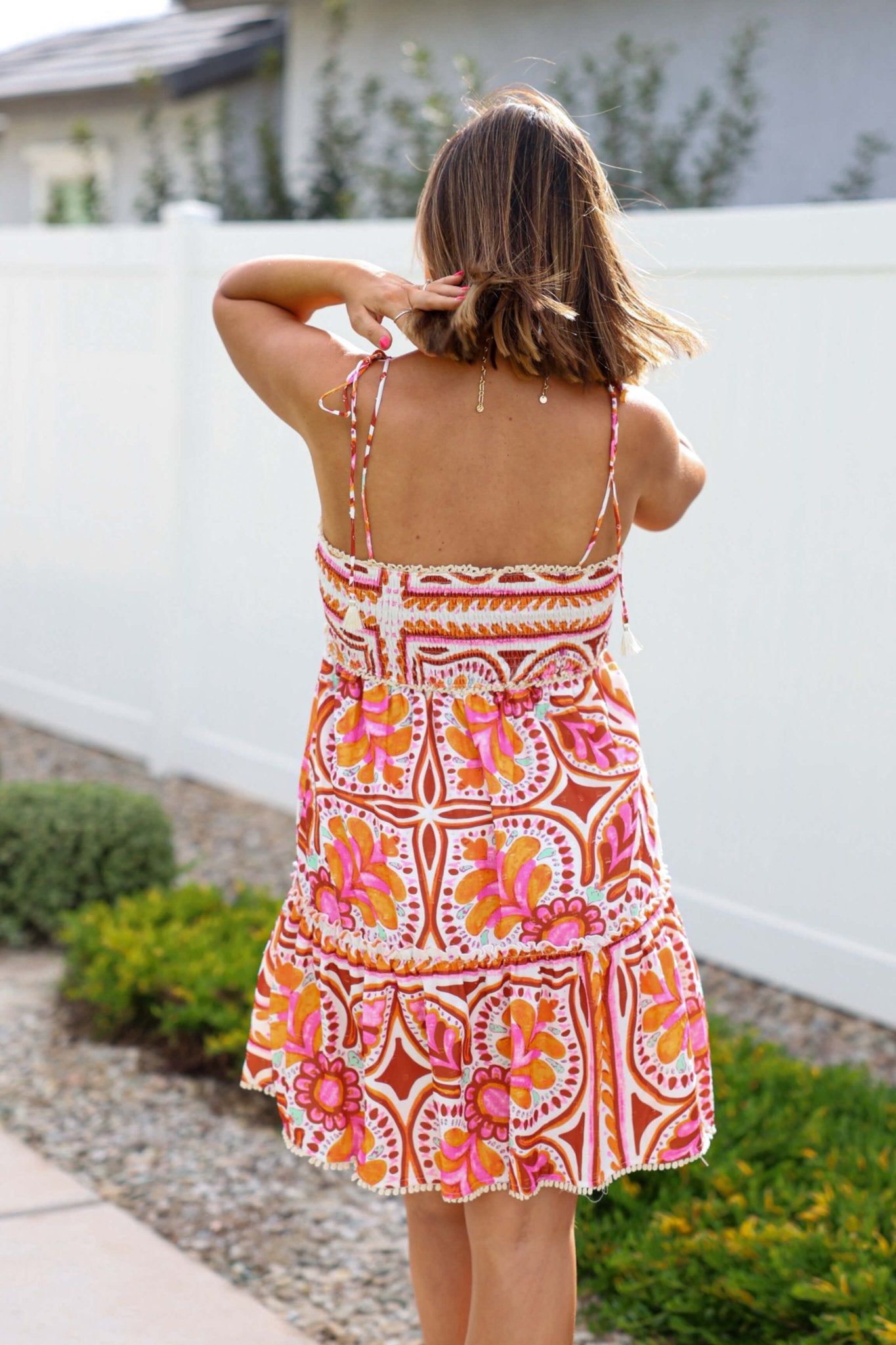 Woman outside by a white fence wearing a Pink Multi Floral Print Smock Tiered Mini Dress with tassel-tie straps.