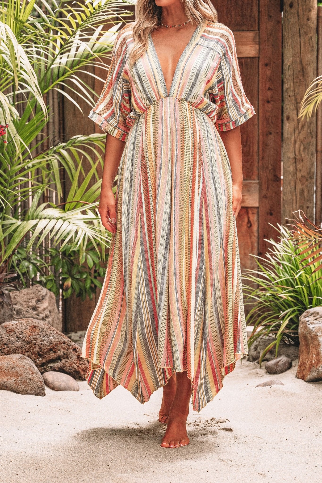 A woman wears the Pink Multi Stripe Kimono Sleeve Midi Dress - FINAL SALE, standing barefoot on sand near tropical plants and wooden fencing.