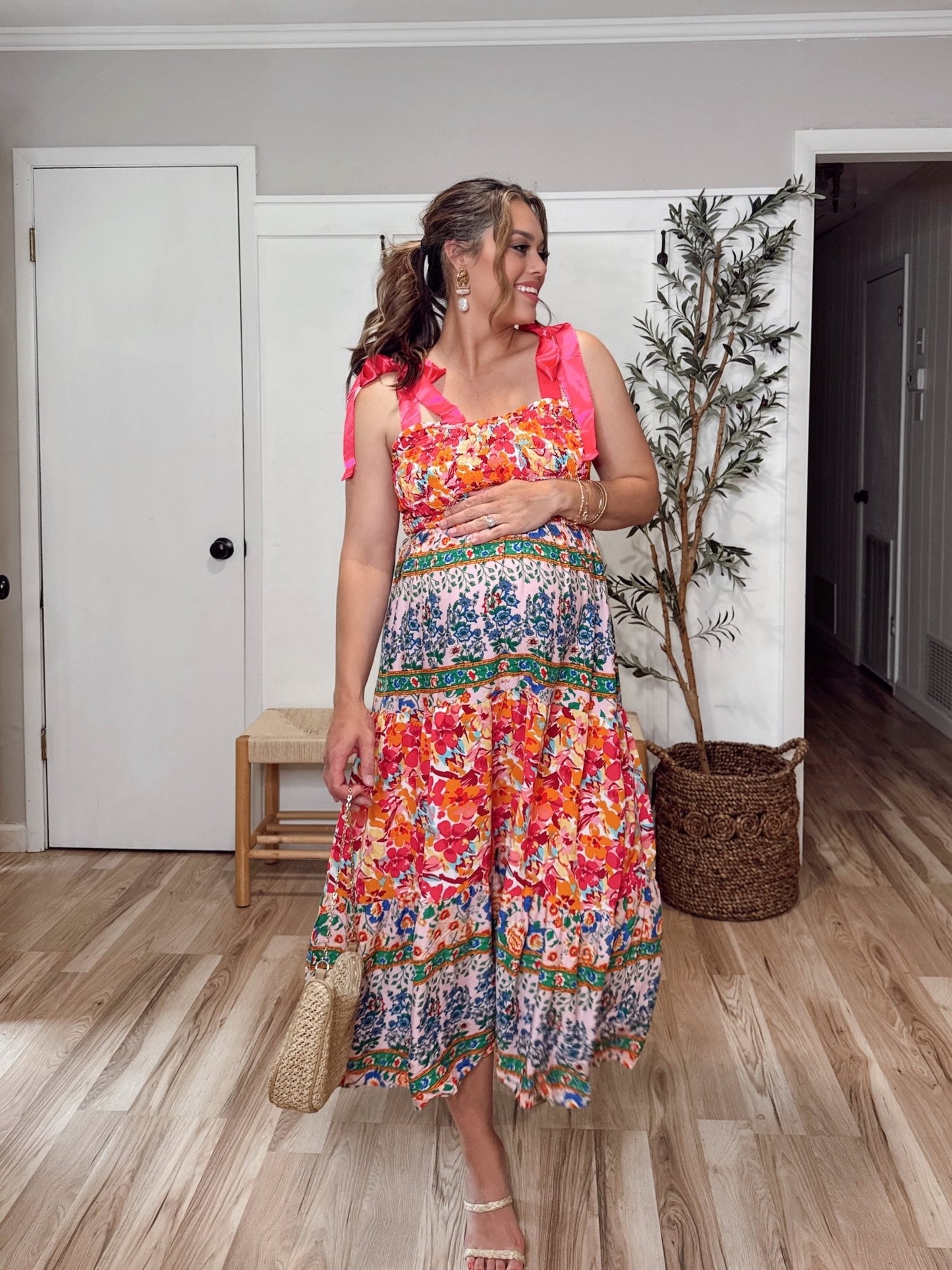 Smiling woman in the Pink Petal Kiss Floral Midi Dress - FINAL SALE, holding a woven bag, stands indoors by a potted plant.