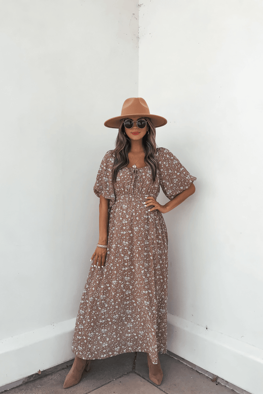 Woman in the Prairie Bloom Maxi Dress - Tan, with a hat, sunglasses, and boots, poses confidently against a white wall.