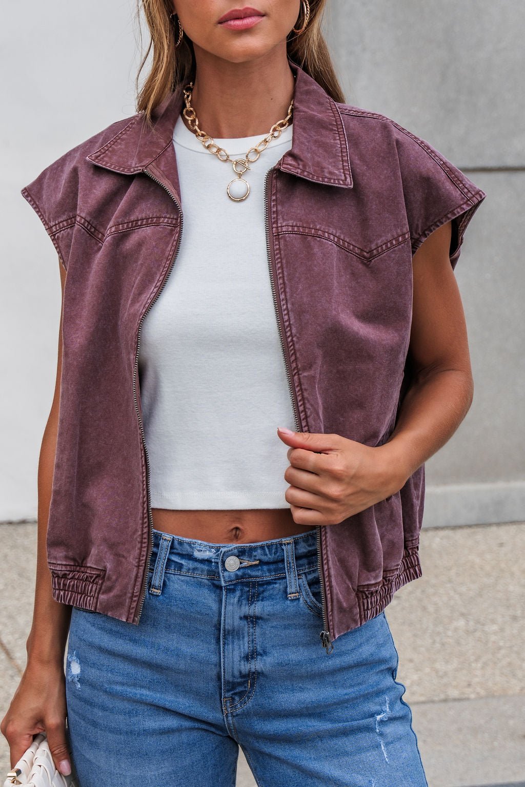 Woman wears Presley Zip Up Collared Vest in mauve over a white crop top and blue jeans, holding the vest open.