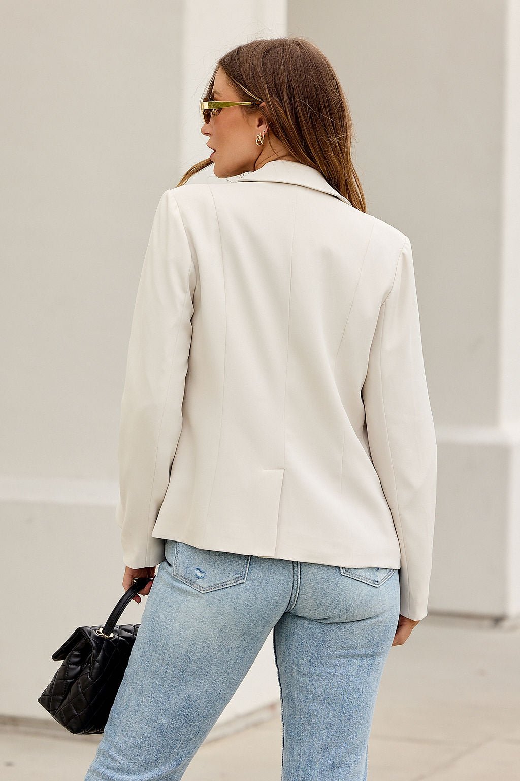A woman in light blue jeans and the Professional Stripe Lined Blazer - Oatmeal holds a black purse, standing by a white wall.