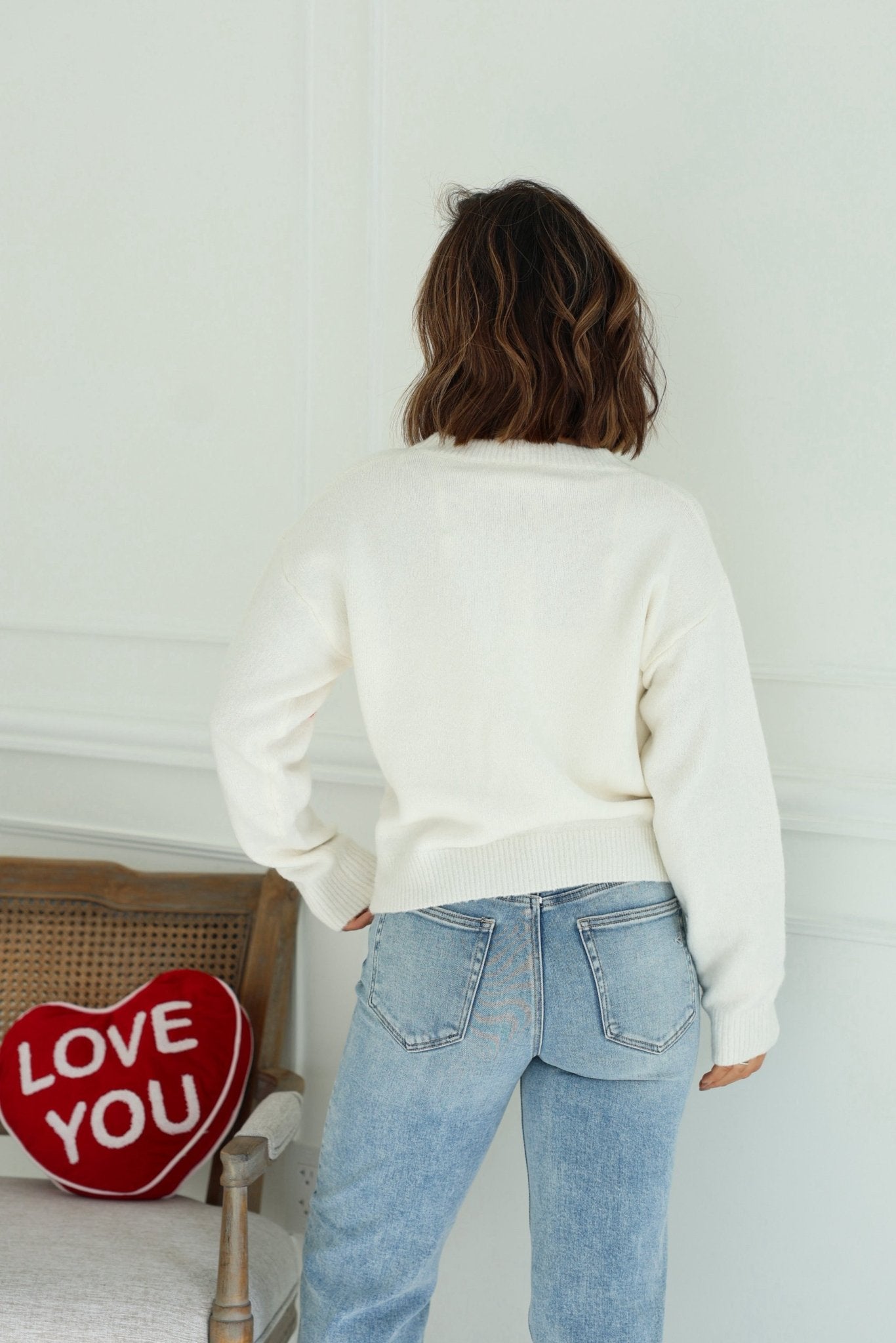 A person wears a Red and White 3D Heart Sweater with blue jeans, standing near a chair with a red heart-shaped LOVE YOU pillow.