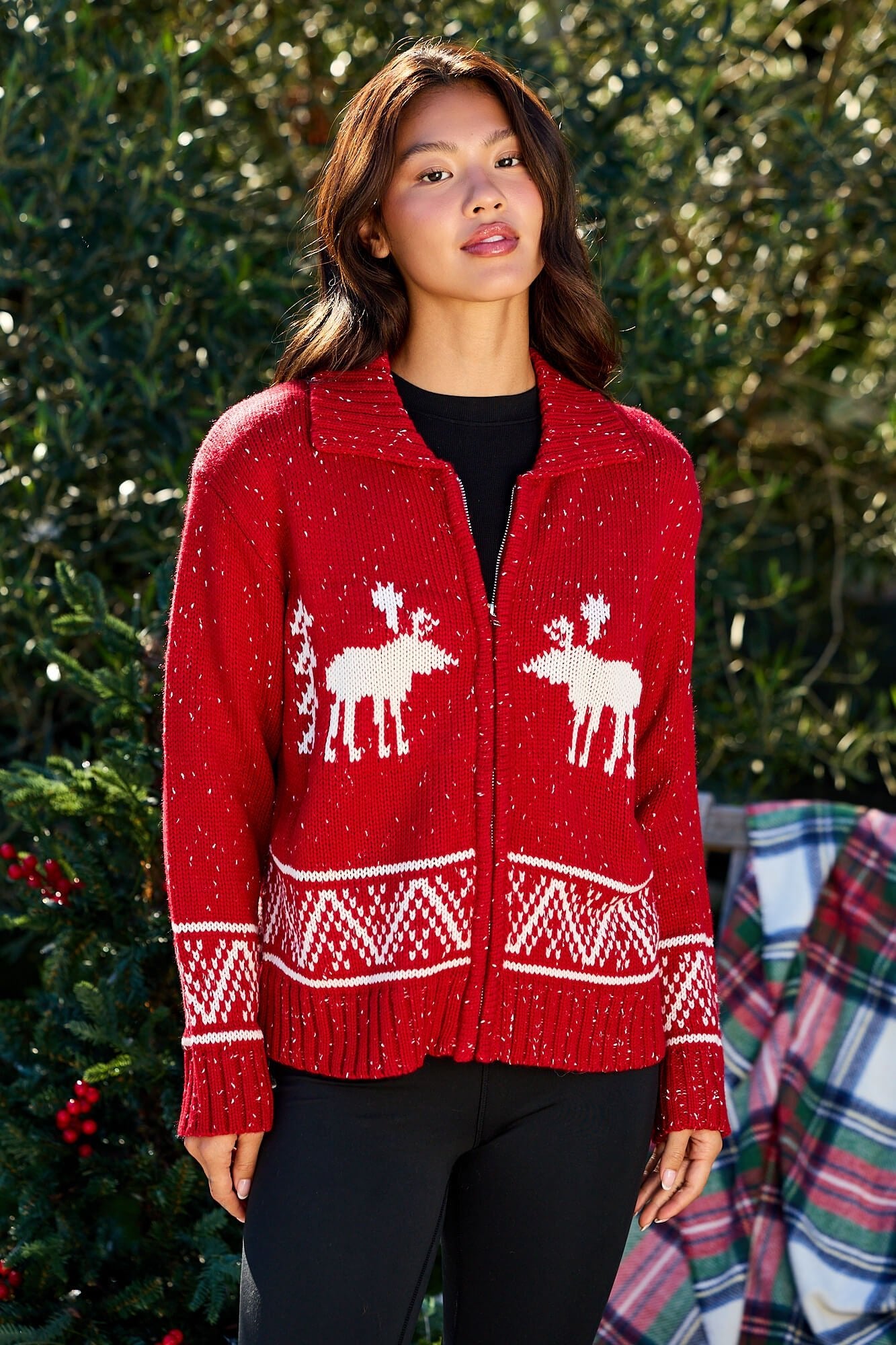 Woman wears a Red and White Aztec Zip Sweater Cardigan outdoors with greenery and a plaid blanket in the background.