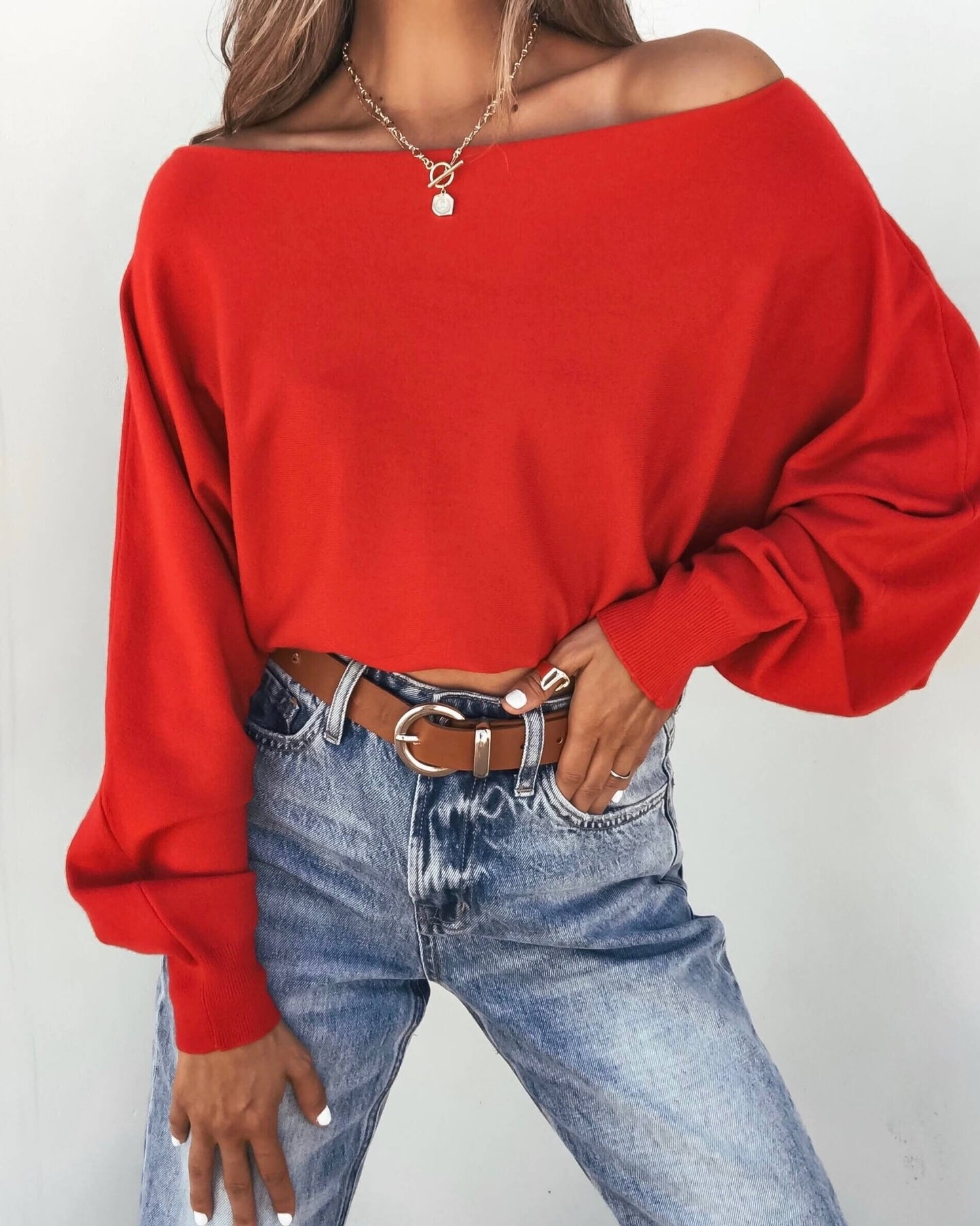 Woman poses in a Red Boatneck Cashmere Sweater, blue jeans, brown belt, necklaces, and rings with one hand on her hip.