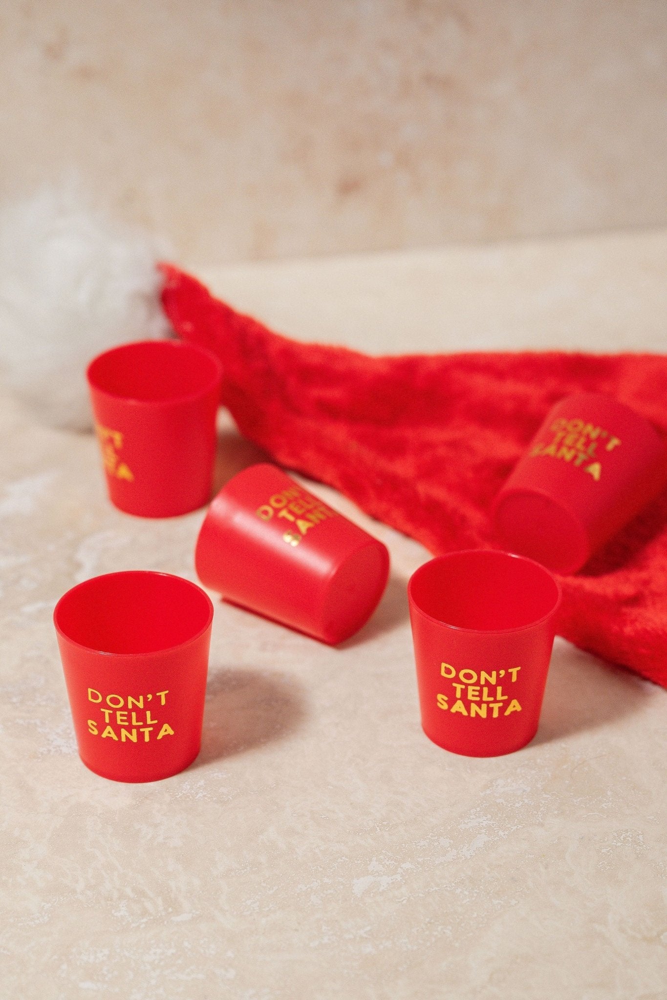 Red Don't Tell Santa Shot Cup Set—five shot cups, ideal stocking stuffers, pictured beside a Santa hat on a light surface.