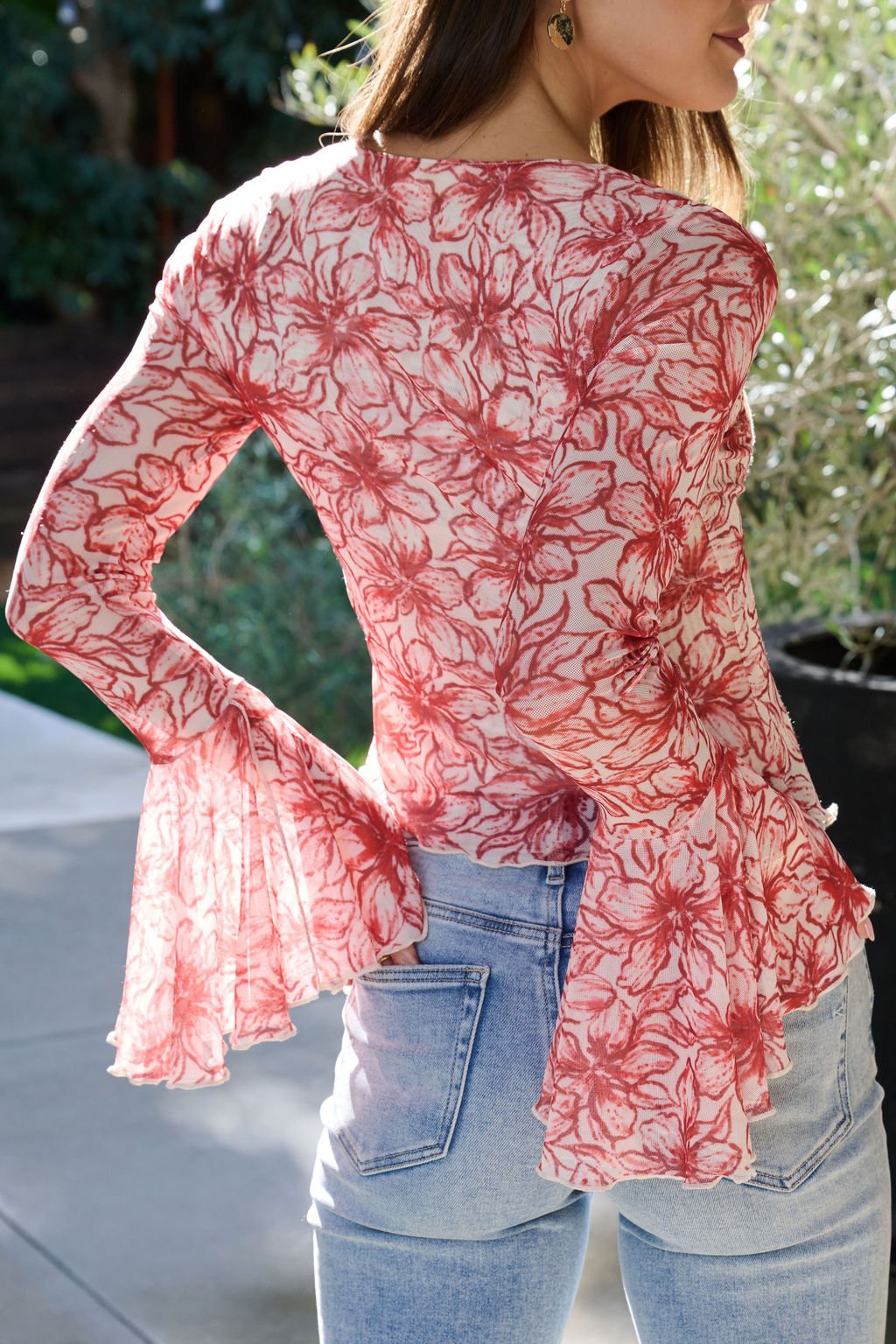 Woman in light blue jeans and a red floral print bell sleeve mesh top, standing outdoors, facing away from the camera.