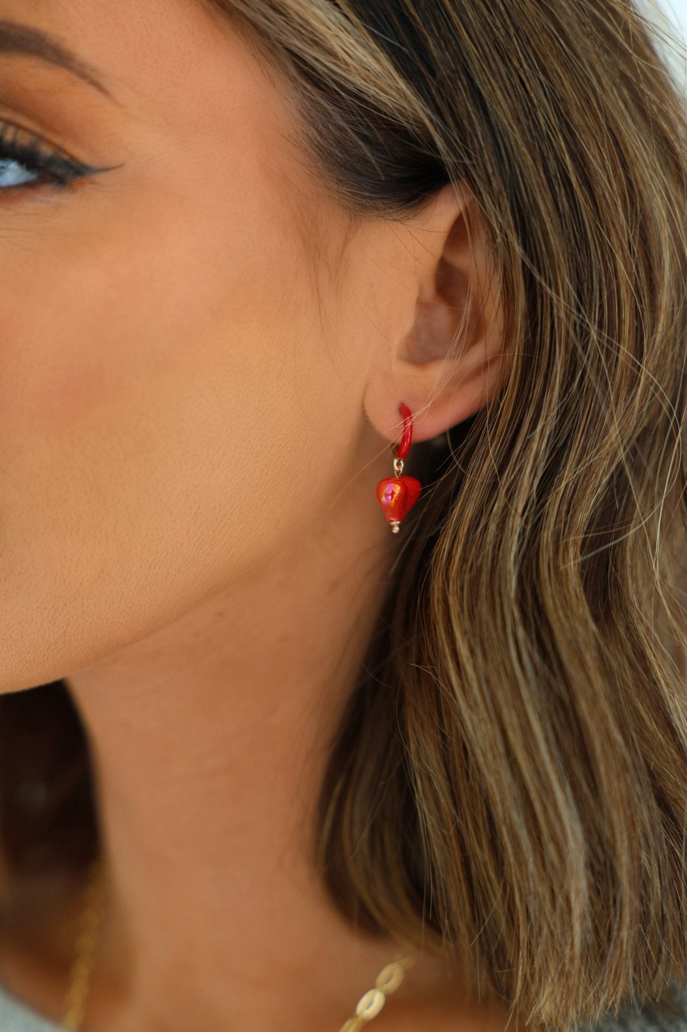 Close-up of a woman's ear wearing Red Heart Charm Hoop Earrings; she has shoulder-length brown hair with blonde highlights.