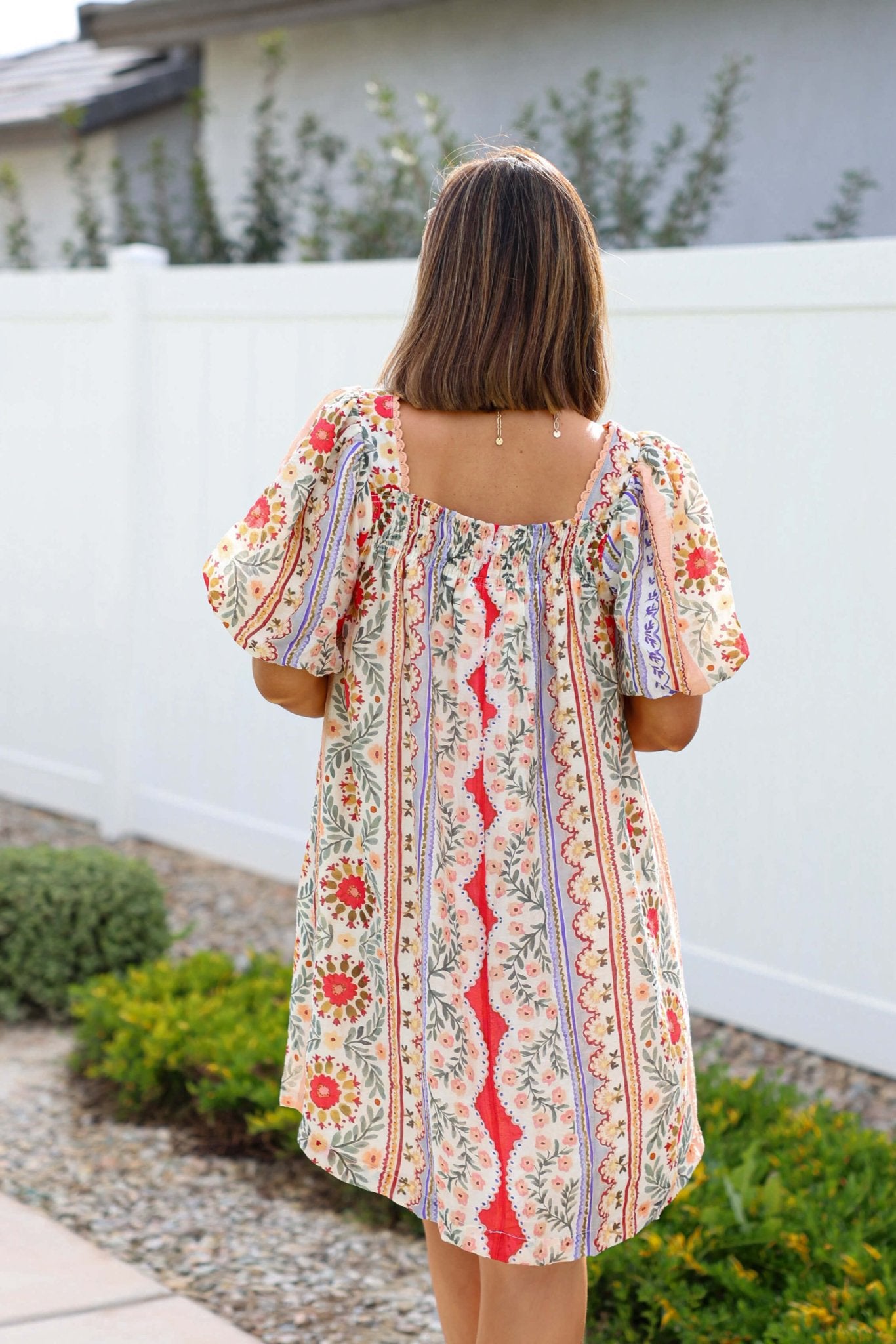 A woman wearing the Red Mix Print Puff Sleeve Mini Dress - FINAL SALE stands outside by a white fence and garden.
