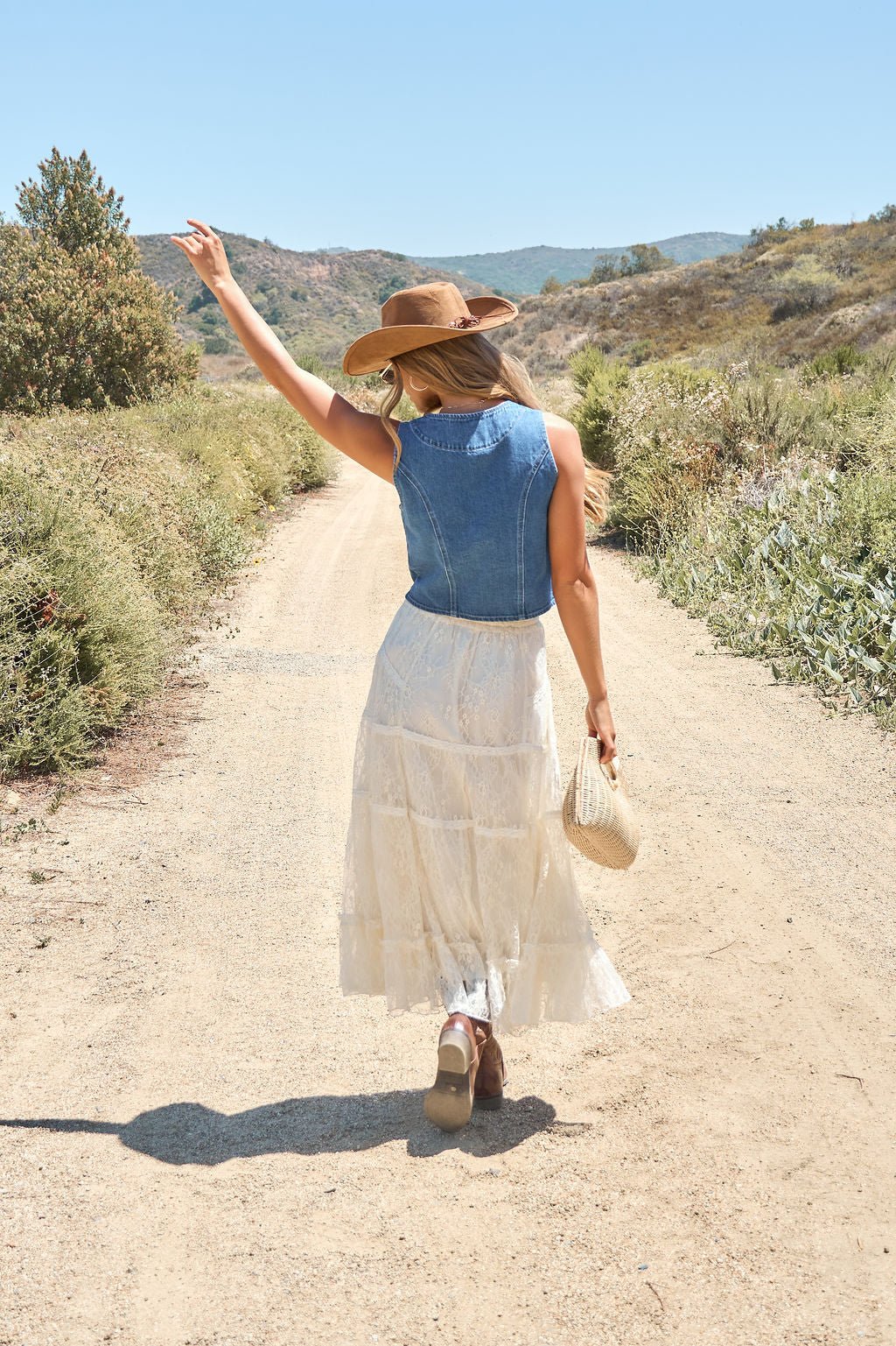 Woman in a denim vest, Rio Lace Tiered Midi Skirt - Cream, and sun hat walks a sunny rural path holding another hat.