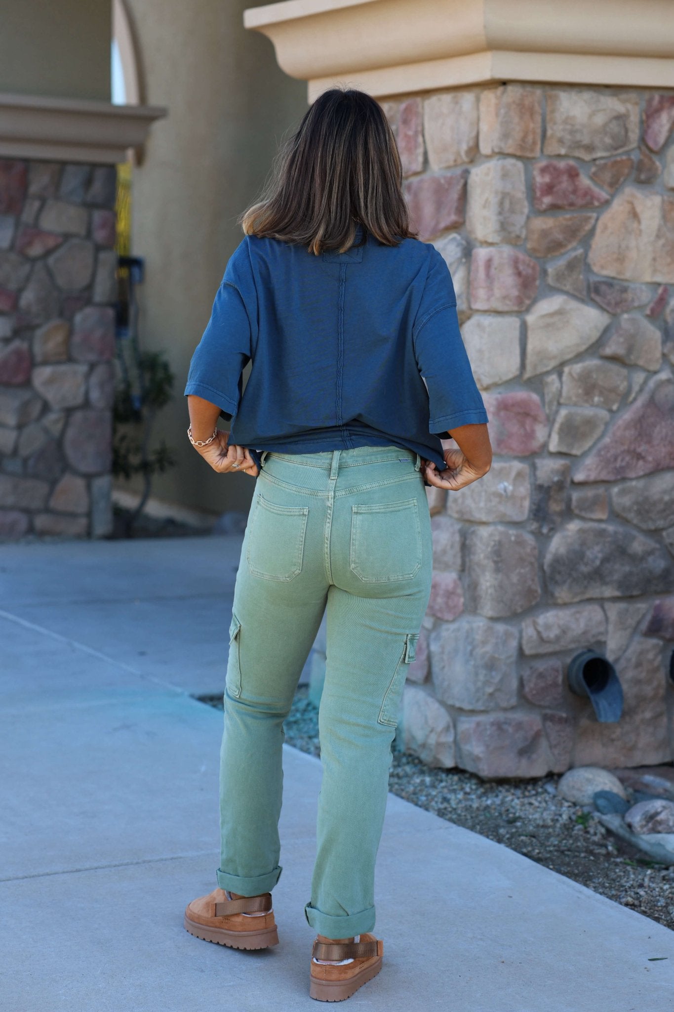 A woman stands outside on a sidewalk, wearing Risen Olive Straight Leg Cargo Pants, a blue top, and brown shoes.