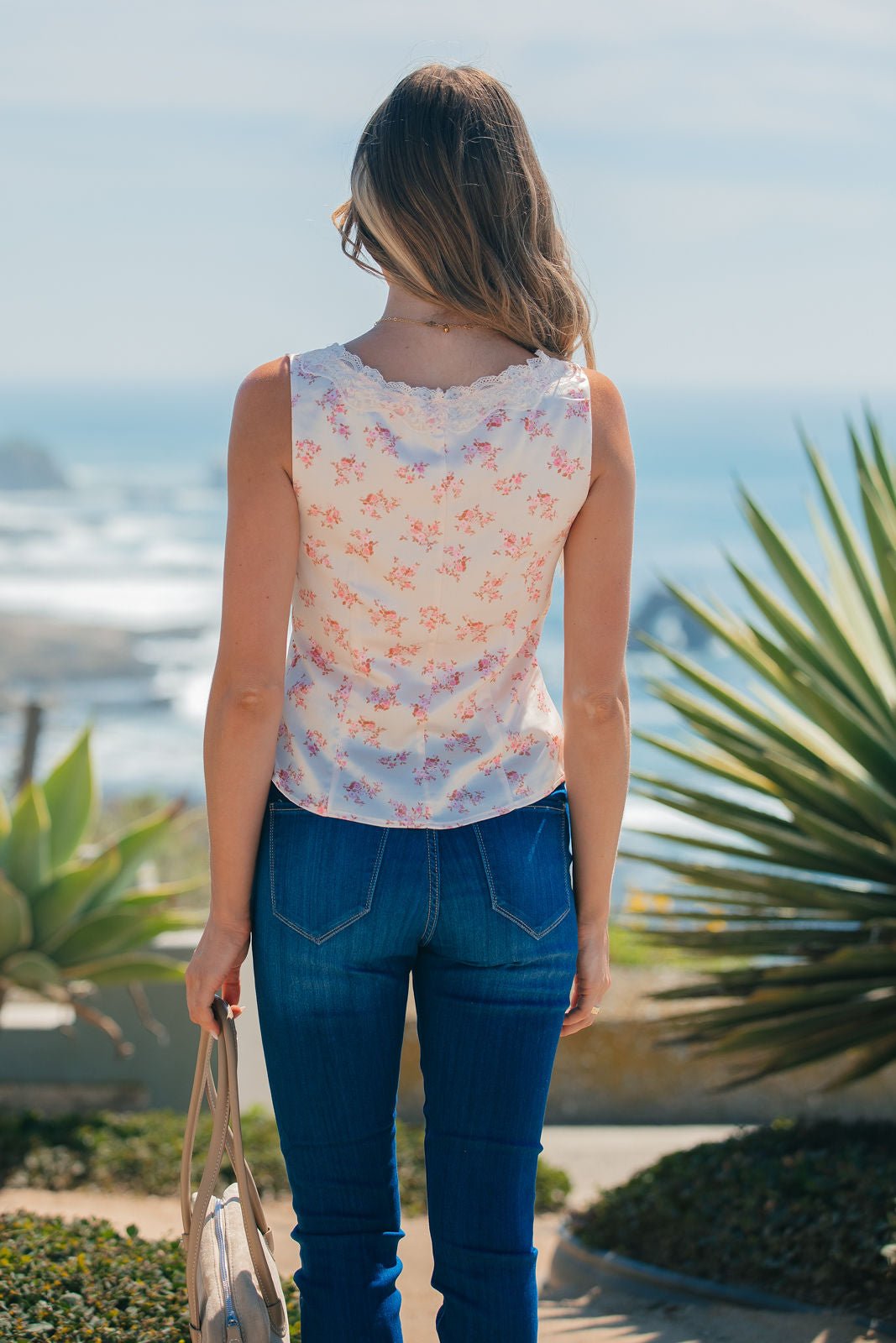 A woman stands outdoors facing the ocean, wearing a Rosy Pink Floral Lace Tank Top and jeans, holding a handbag.