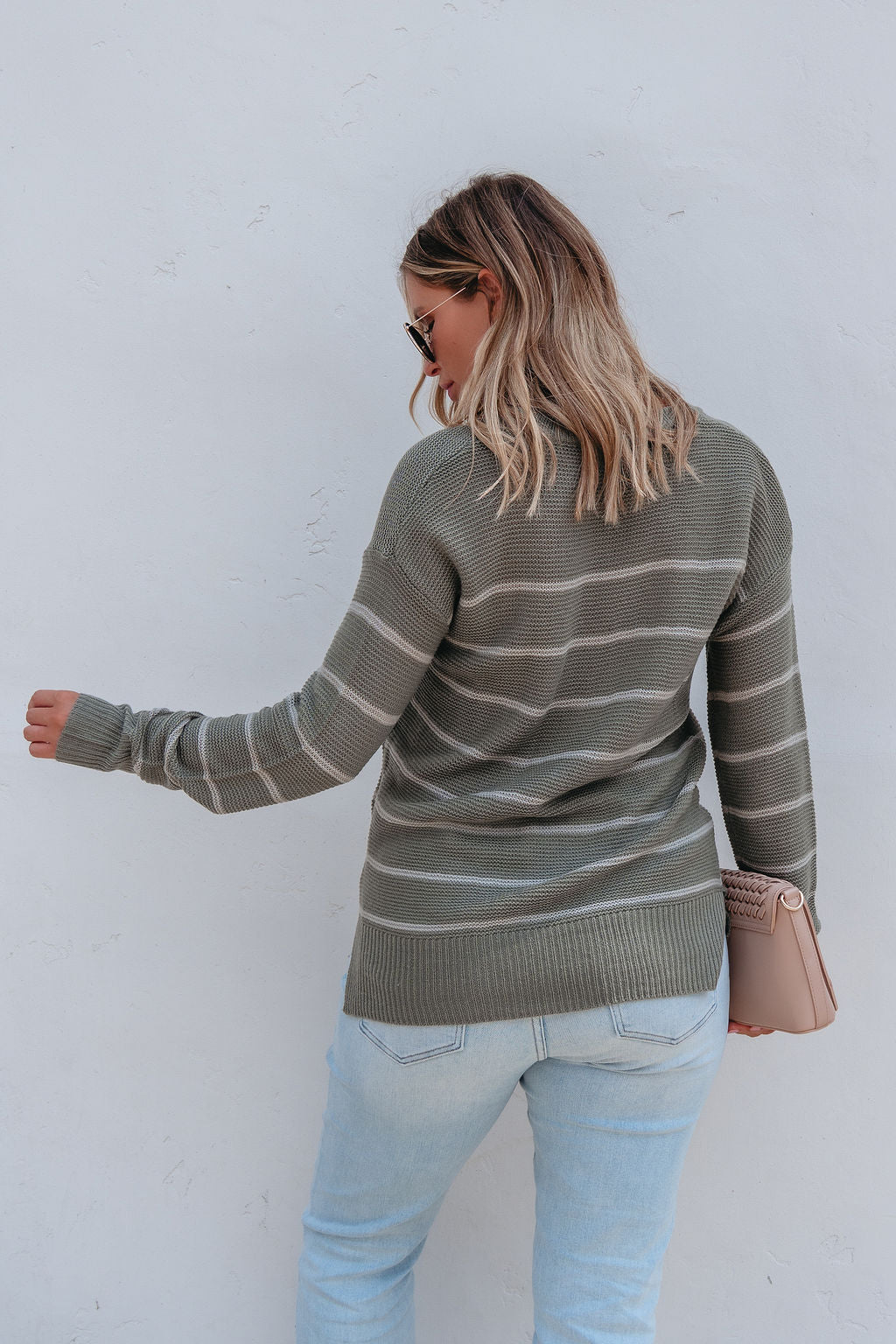A woman faces away wearing the Rowan Olive Striped Tunic Sweater, light jeans, and holding a small pink purse.