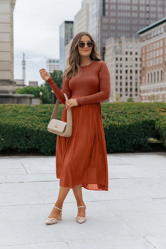 Woman in a Rust Contrast Pleated Midi Dress, sunglasses, and heels holds a beige handbag on the city sidewalk.