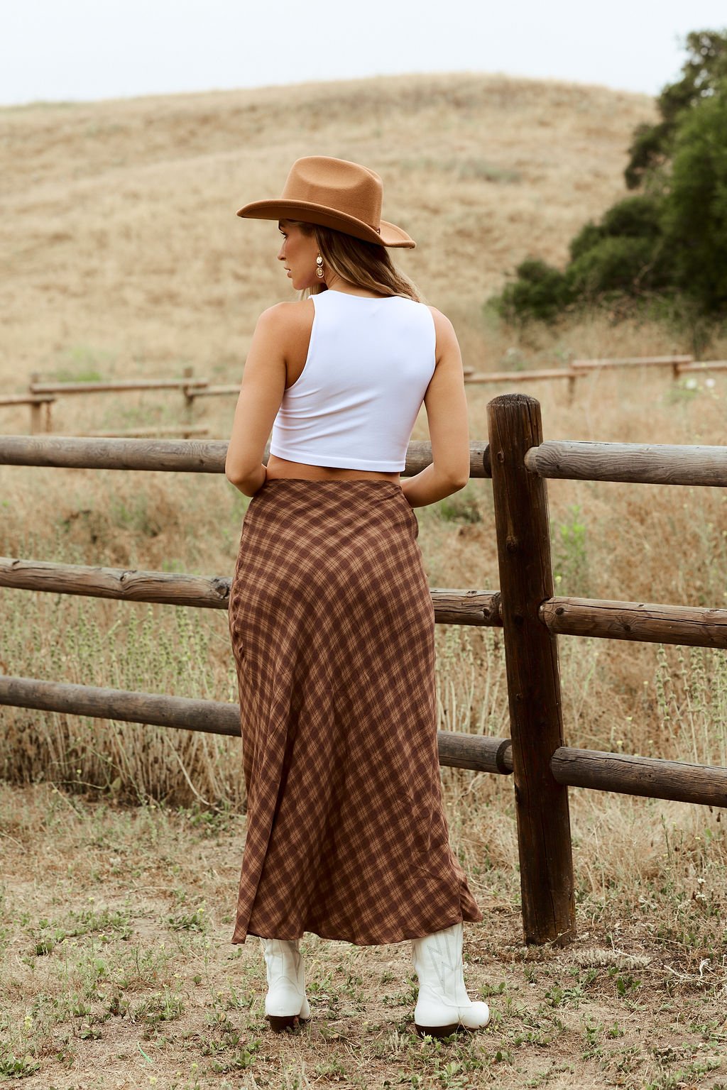 A woman in a cowboy hat and the Saddle Ridge Brown Plaid Maxi Skirt - FINAL SALE with a high waist and A-line silhouette.