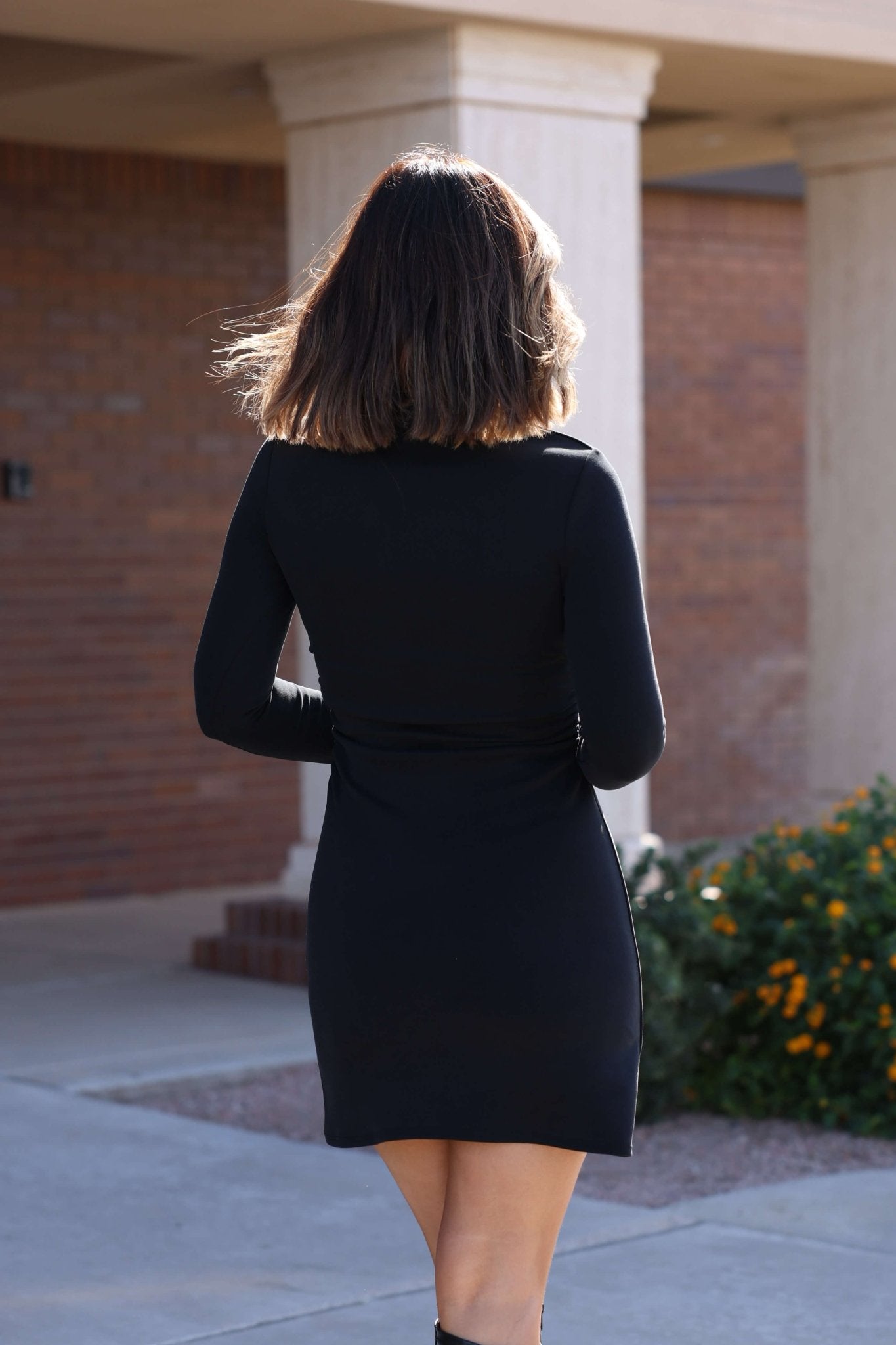A woman in a Sadie and Sage Black Ruched Mini Dress stands outside by yellow flowers and a building.