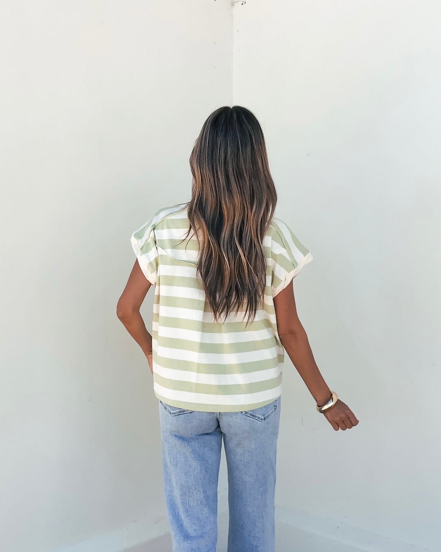 A woman in the Sage and Ivory Striped Button Up Shirt and jeans stands with her back to the camera, facing a white wall.