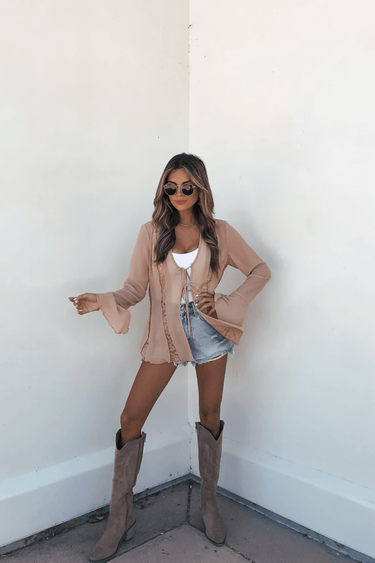 Woman in sunglasses wears a Sand Bell Sleeve Sheer Lace Top, denim shorts, and tan knee-high boots against a white wall.