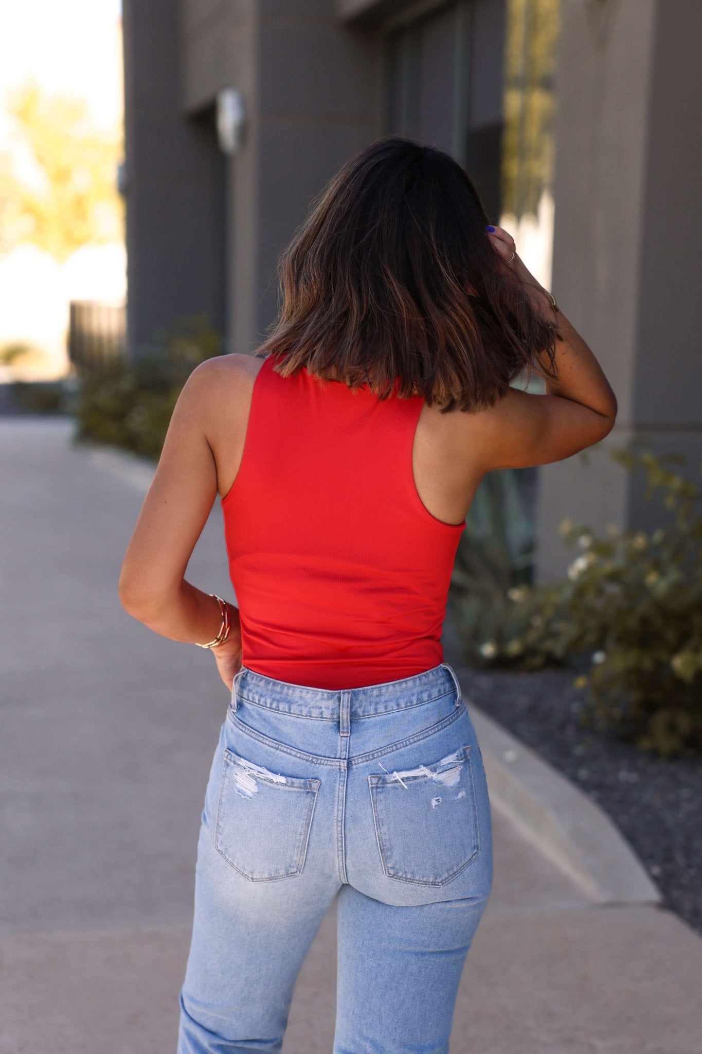 A person with shoulder-length brown hair wears the Sangria Nights Red Tank Bodysuit - DOORBUSTER and light blue jeans outdoors, facing away.