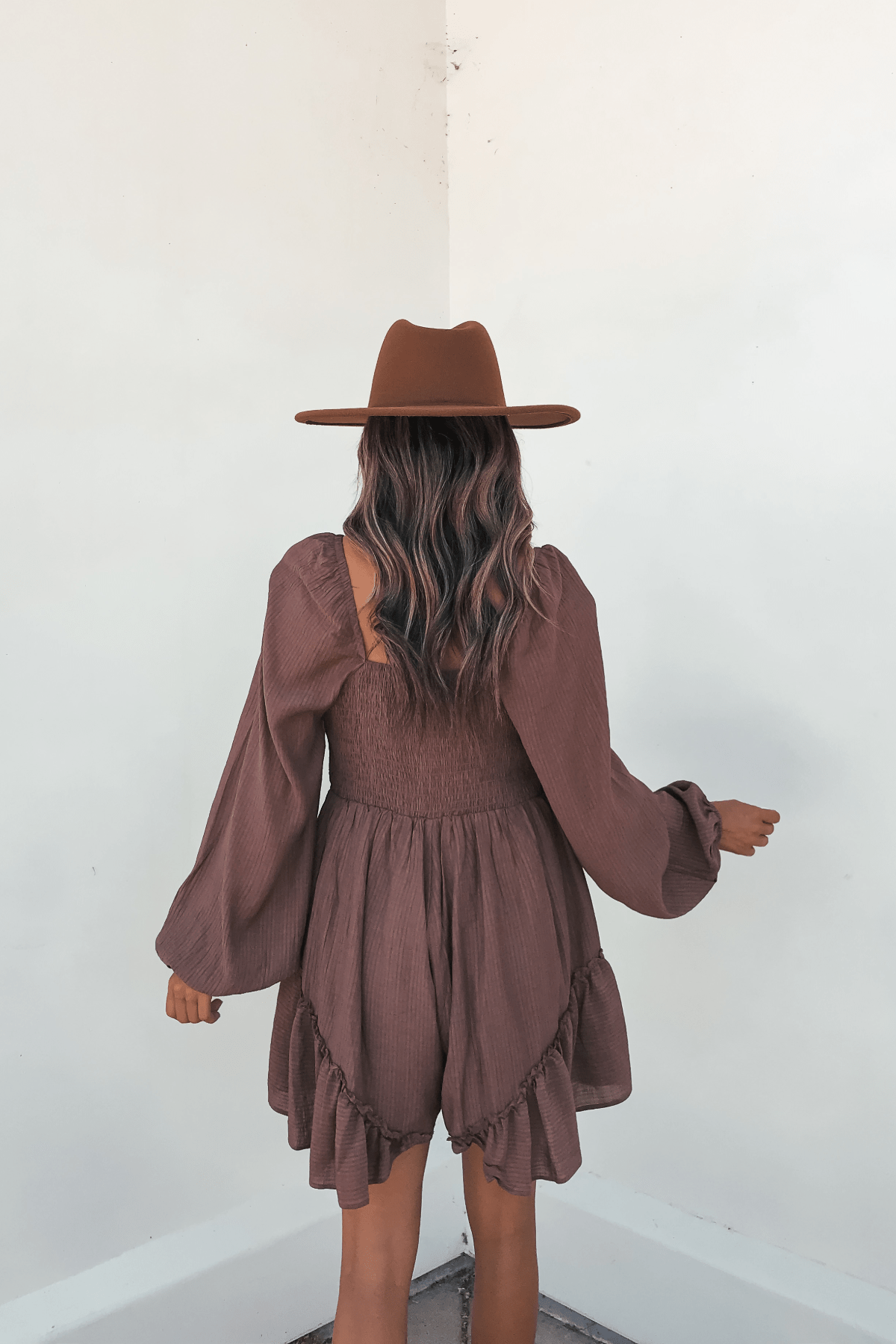 Woman with wavy hair wears the Shay Mocha Ruffled Sweetheart Romper, standing back to camera against a white wall.