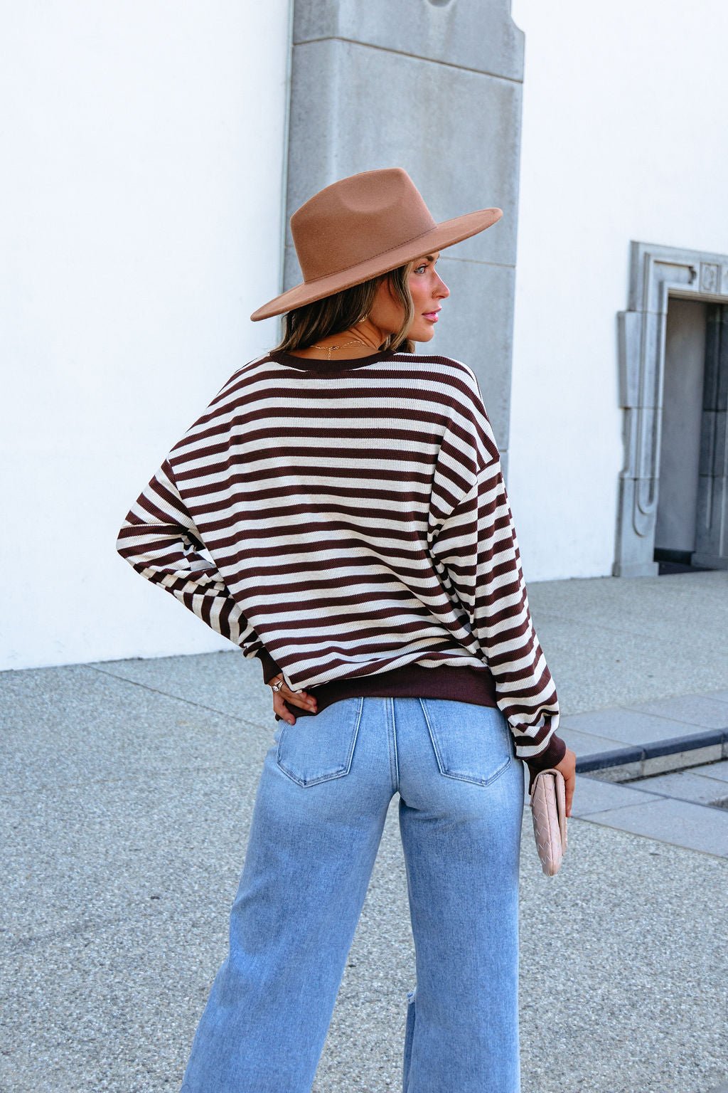 Woman models SHE AND SKY ST1813 brown striped top outdoors, paired with a brown hat and blue jeans, facing away from the camera.