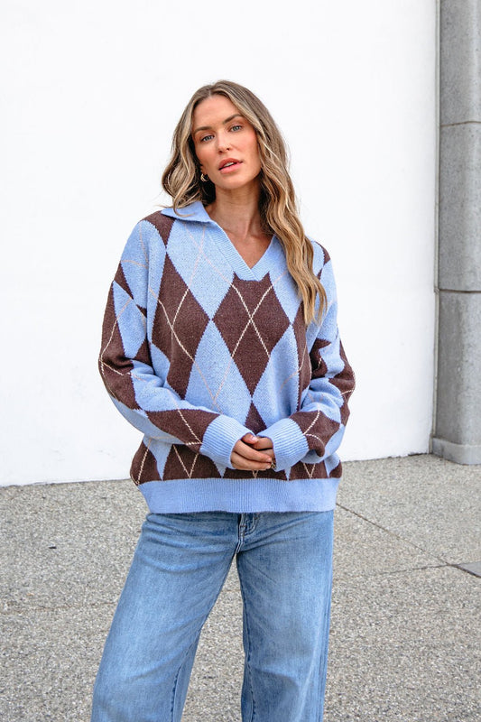 Woman in a Sky Blue Argyle Ribbed Sweater and jeans stands outside by a white wall and gray stone column.