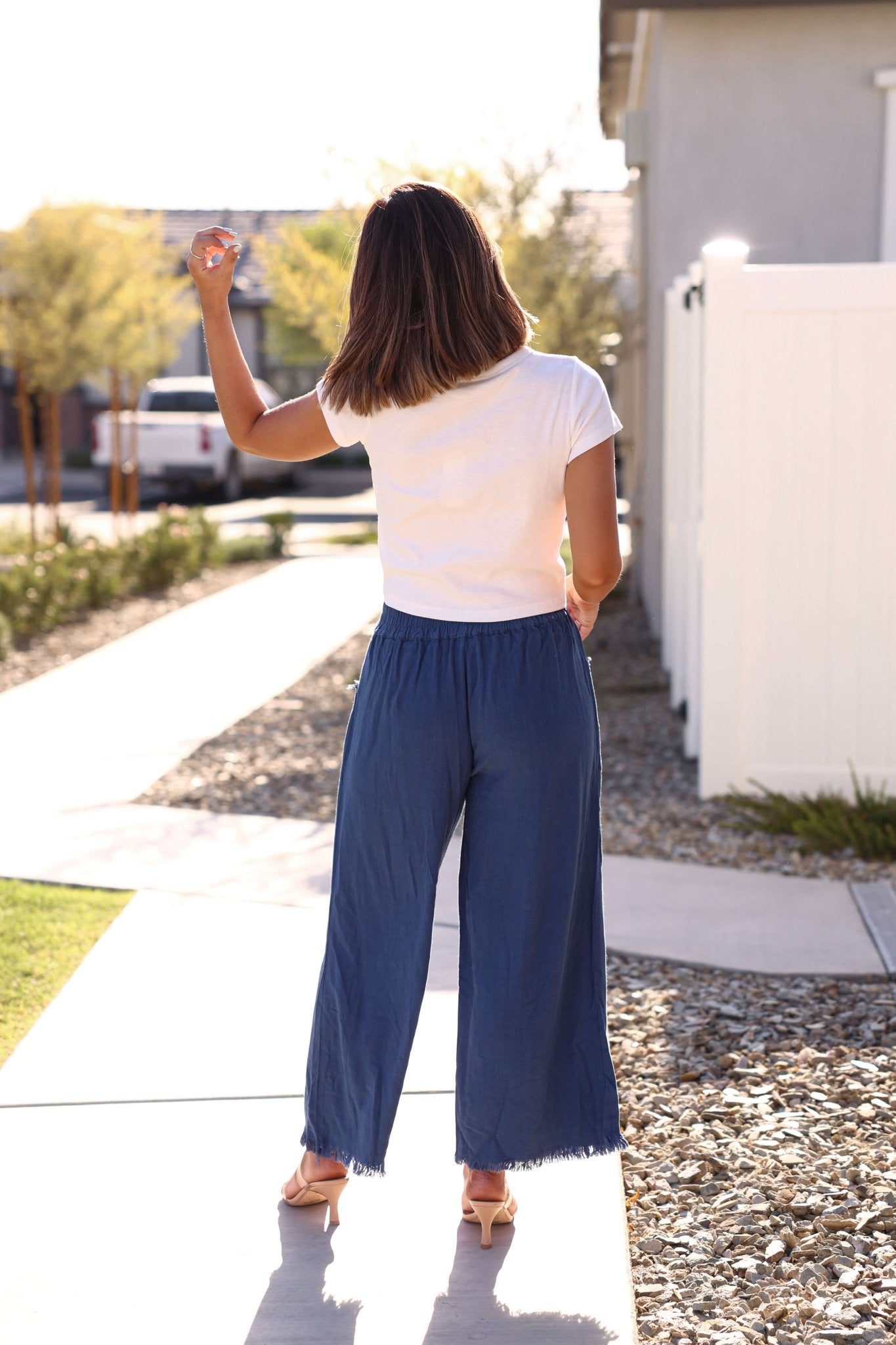 A woman stands on a sunny sidewalk in a white top and Slate Blue Frayed Linen Wide Leg Pants, facing away from the camera.