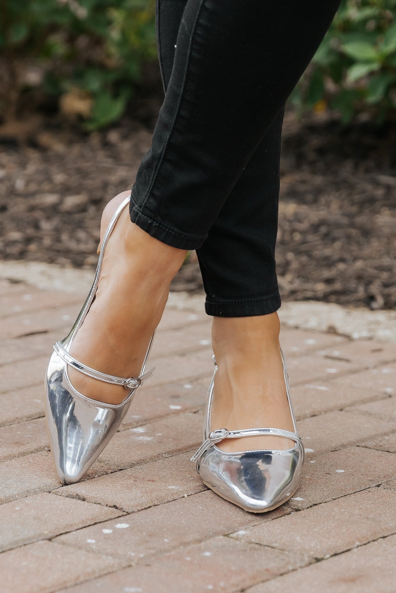 A person wearing Sleek Silver Buckle Detail Slingback Heels - DOORBUSTER and black pants, standing on a brick path.