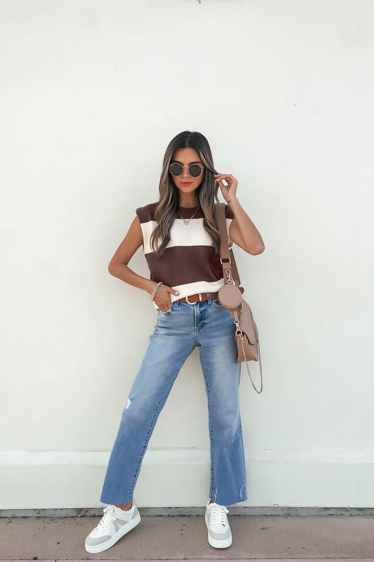 Woman in sunglasses wears a sleeveless brown and cream striped sweater, blue jeans, white sneakers, and a beige crossbody bag.